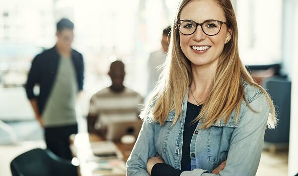 Confident young businesswoman smiling while standing with her arms crossed in a modern office with colleagues working in the background